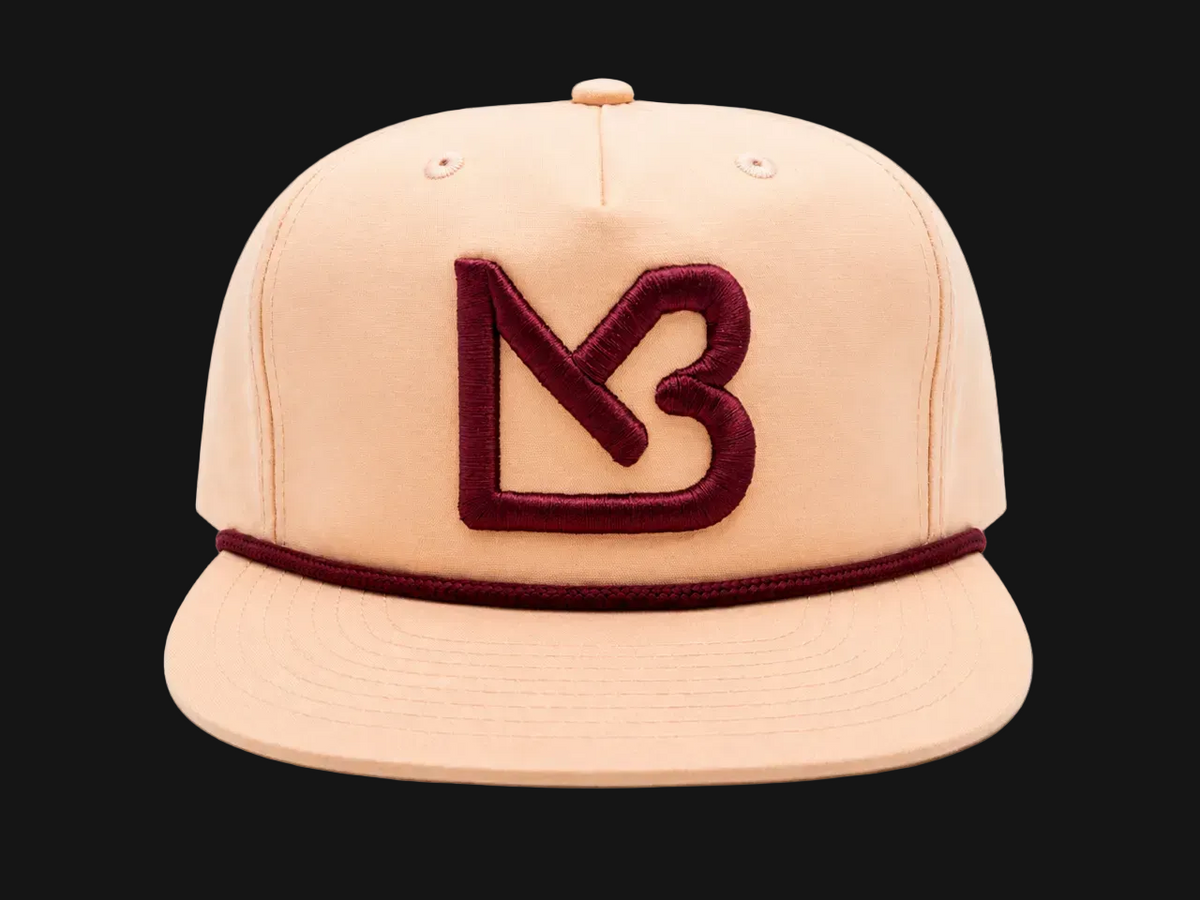 A structured flat-brim cap in a light peach-beige color, featuring a prominent dark burgundy embroidered 'MB' logo on the front and a matching burgundy rope detail around the crown.